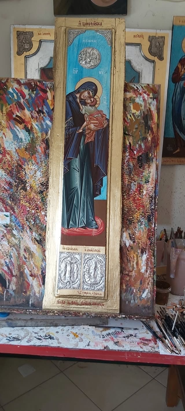 Orthodox religious icon depicting a saint holding an infant, with ornate wooden frame and decorative floral tapestries displayed in an artists studio