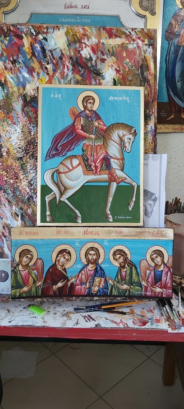 Orthodox religious icon artwork in progress showing saint on horseback and five figures below, displayed on artists work table with paint palette and brushes