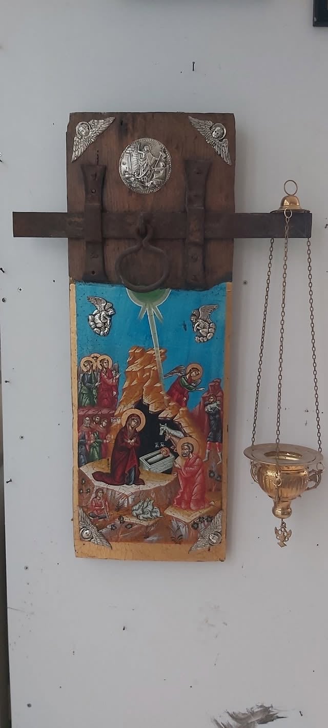 Wooden religious icon panel with nativity scene mounted on white wall next to hanging brass ceremonial vessel.