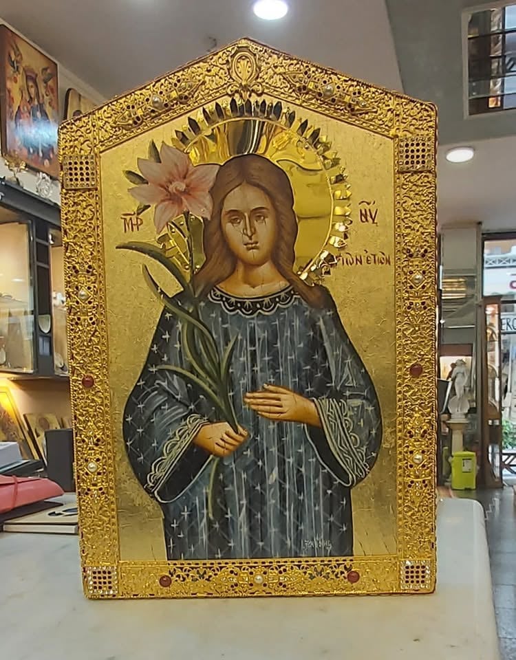 Orthodox Christian icon with gold leaf border depicting a figure in dark robes with a golden halo holding a flower, displayed in an indoor setting