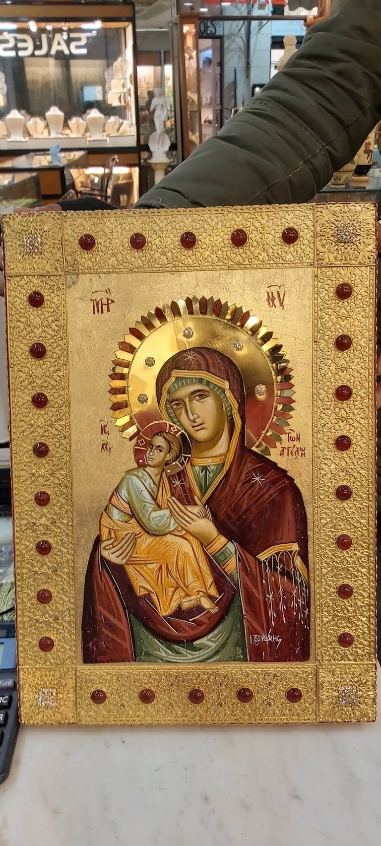 Religious icon depicting Mary holding baby Jesus, painted on gold leaf with jeweled border, displayed in jewelry store.