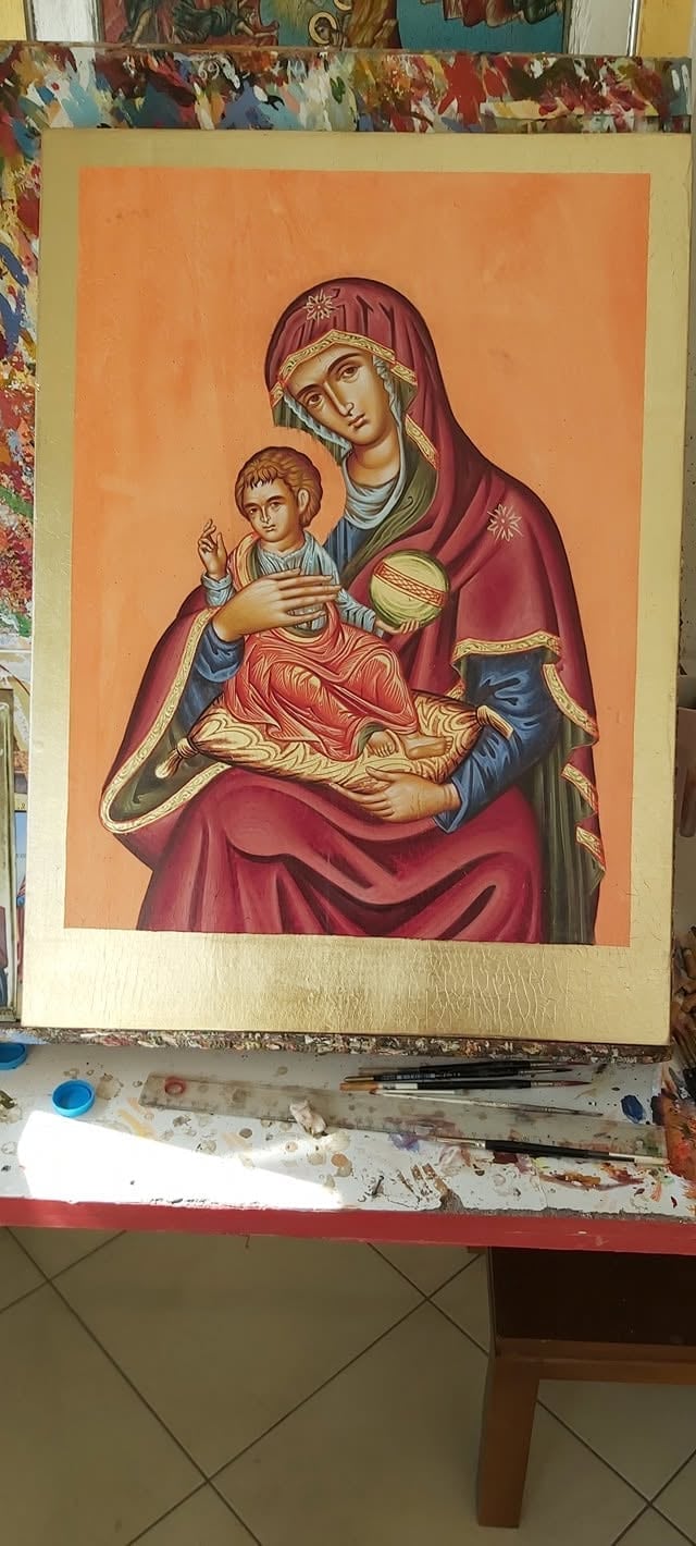 Religious icon painting of Madonna and Child on orange background, displayed on artists table with art supplies