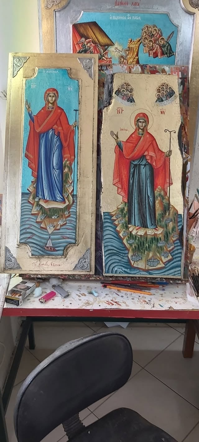 Religious icon paintings displayed on a red table, featuring Orthodox saints in traditional robes with blue water scenes below