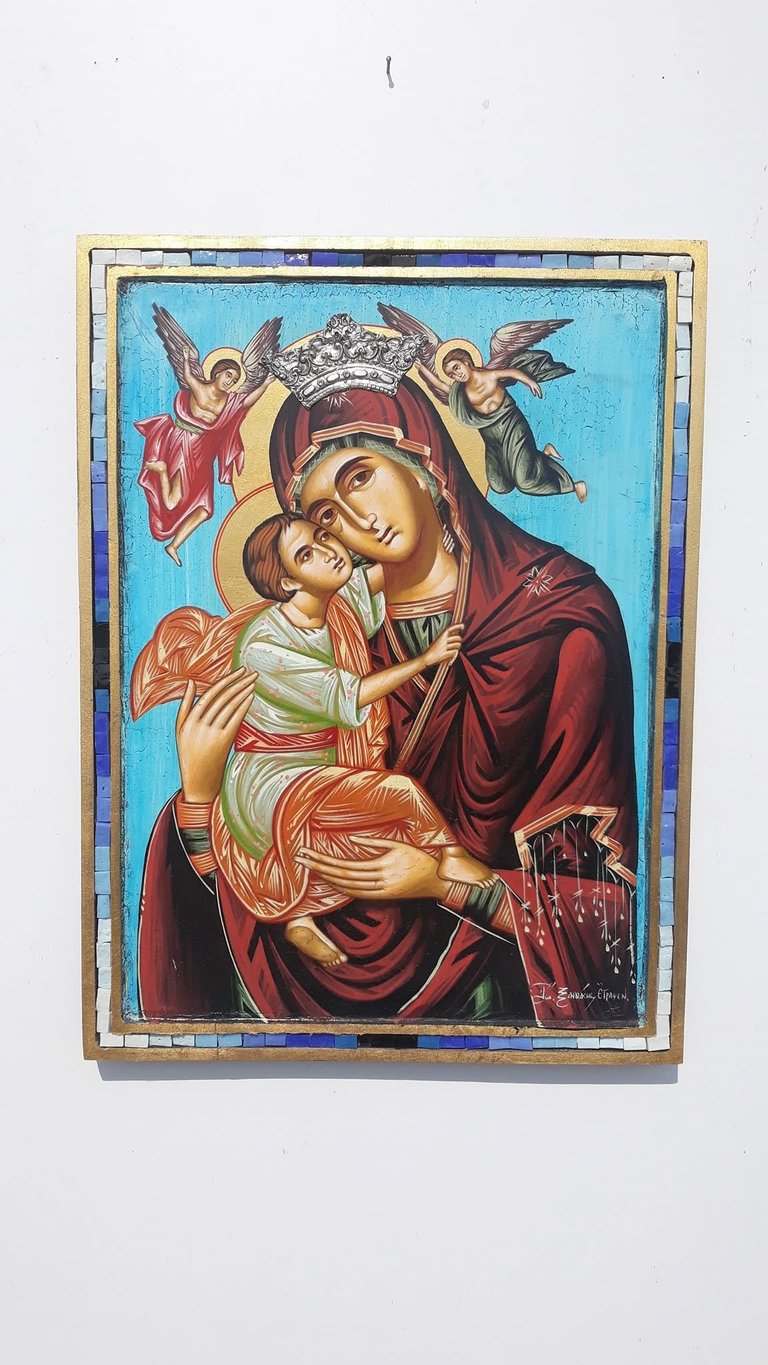 Orthodox religious icon depicting Virgin Mary holding baby Jesus, with golden halos and angels above, framed in decorative border with gold and blue trim