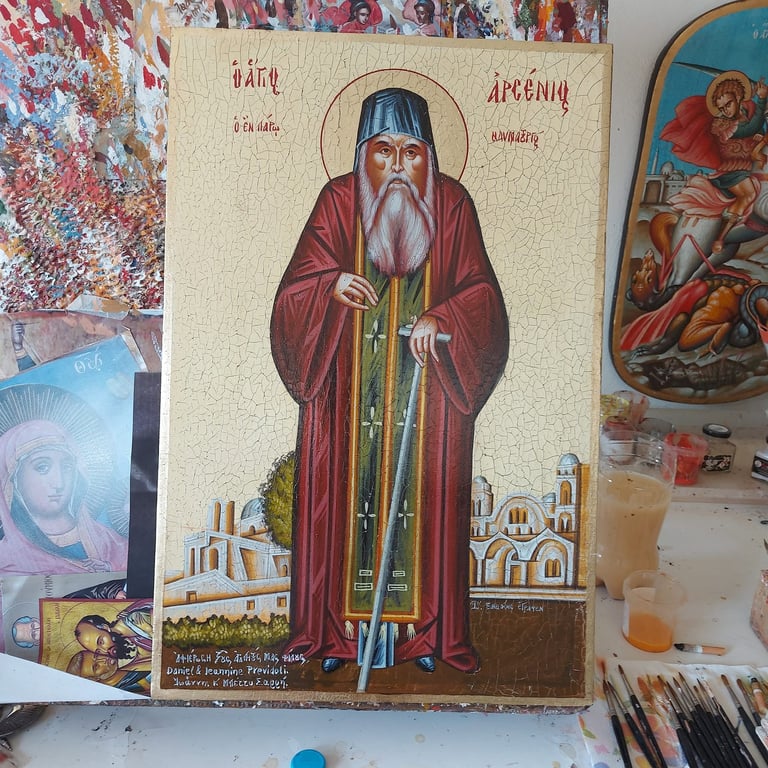 Orthodox religious icon depicting a bearded saint in maroon robes holding a staff, surrounded by monastery buildings at base