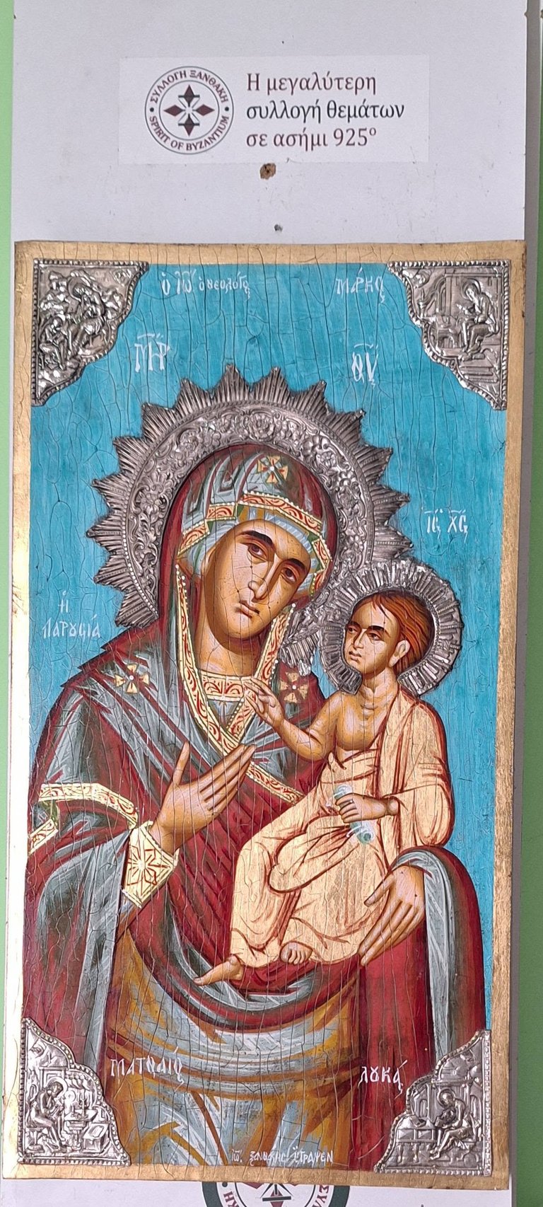 Orthodox icon depicting Virgin Mary in red robes holding Christ Child, with ornate silver halos and blue background, featuring Greek text
