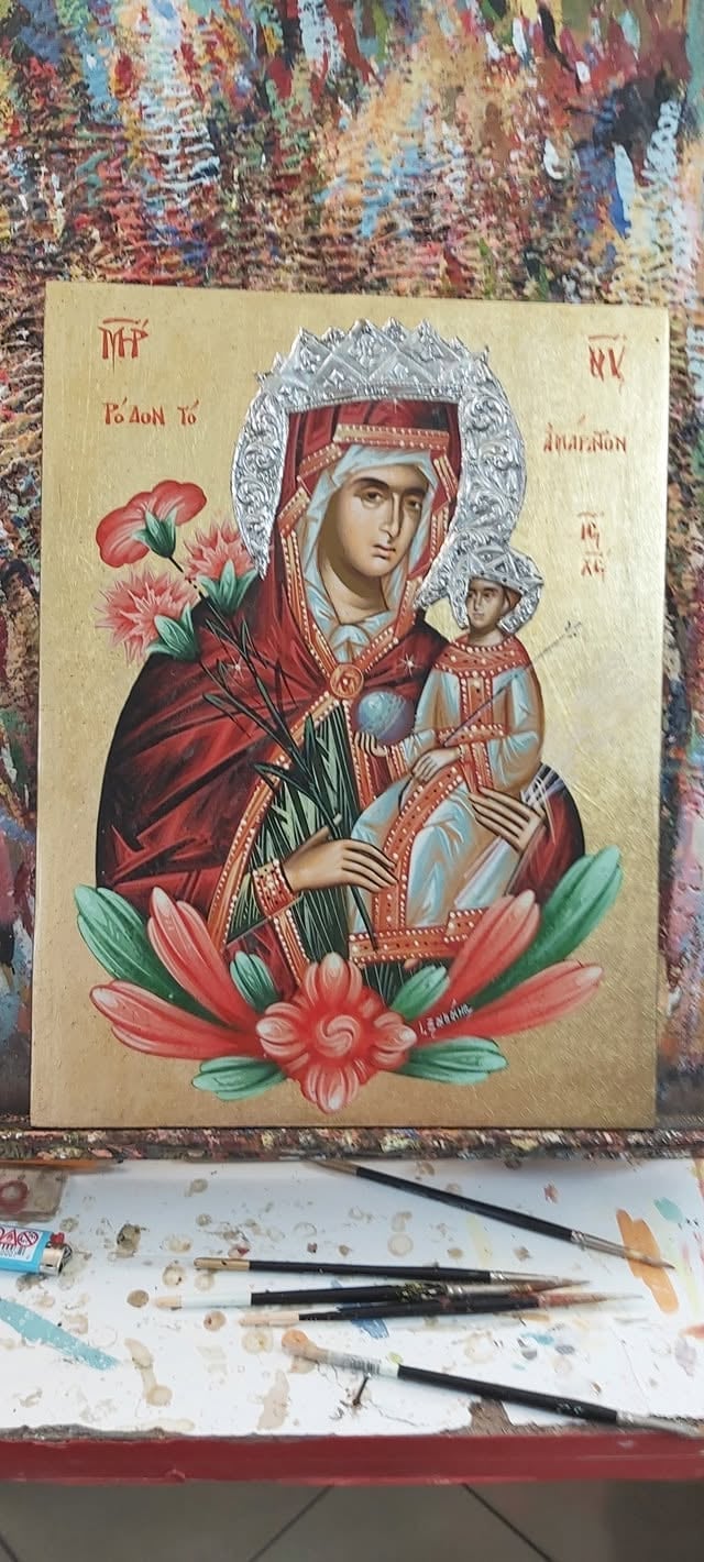 Hand-painted Orthodox religious icon of Virgin Mary with Child Jesus, decorated with pink flowers and gold background, work in progress on artists desk.