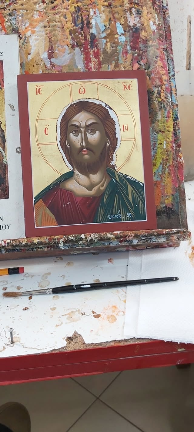 Orthodox Christian icon of Jesus Christ with a gold halo on a red wood panel, displayed in an artists studio with paintbrushes and colorful artwork in background
