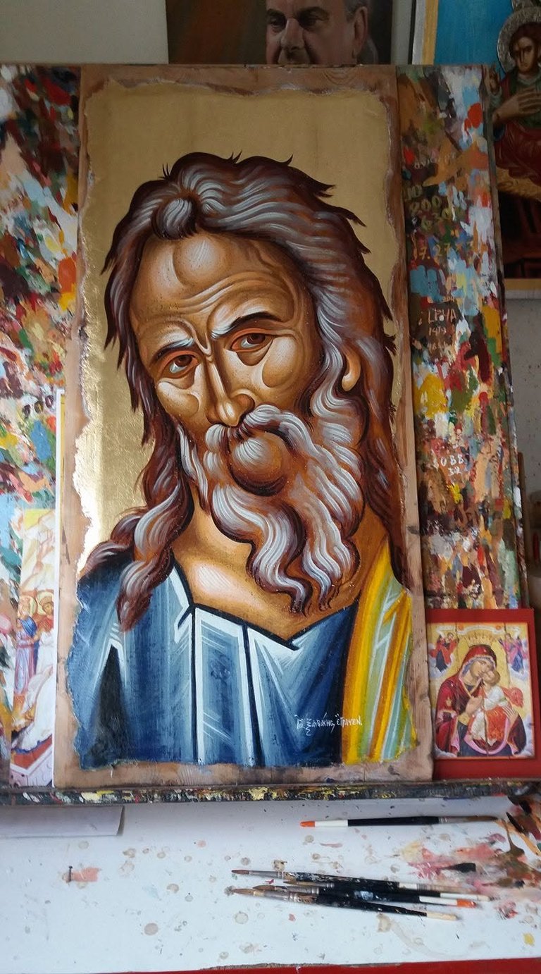 Religious icon painting of a bearded man in blue and gold robes, displayed in an artists workshop with colorful religious artwork and paintbrushes visible