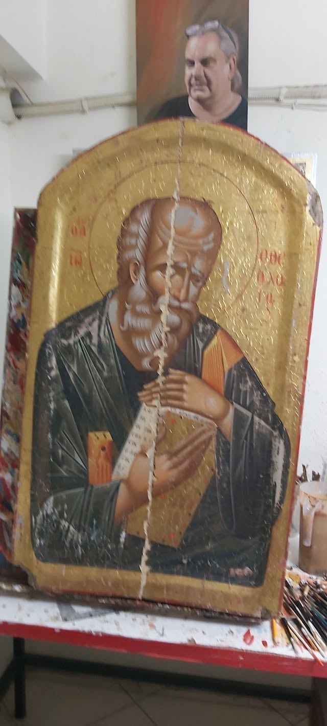 Religious icon artwork on wooden panel with gold background depicting a bearded saint figure holding a cross, displayed on table indoors