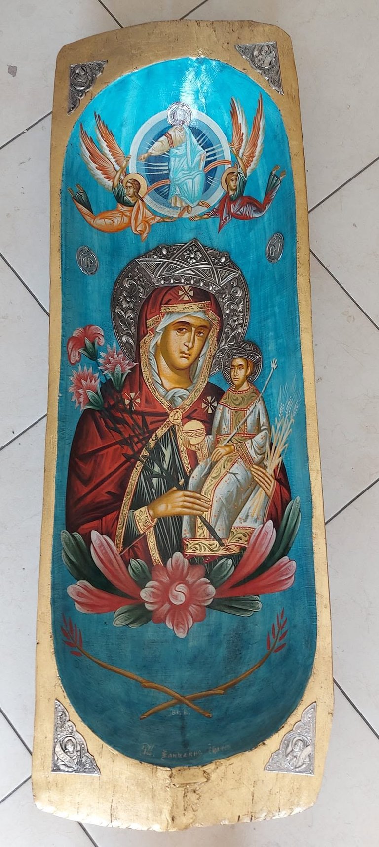 Orthodox religious icon on wooden panel featuring Virgin Mary with child Jesus, angels above, ornamental frame, and pink flowers on turquoise background